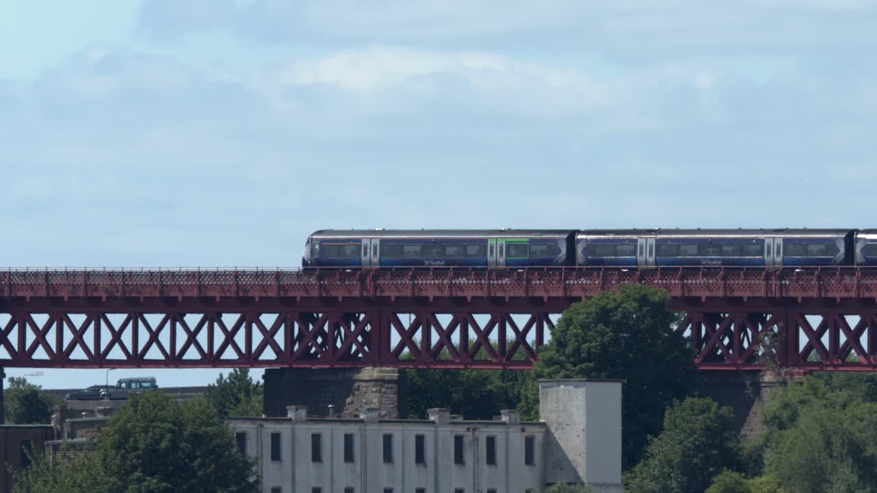 Passenger train travels across iconic red iron bridge in daylight, wide static shot, clear weather