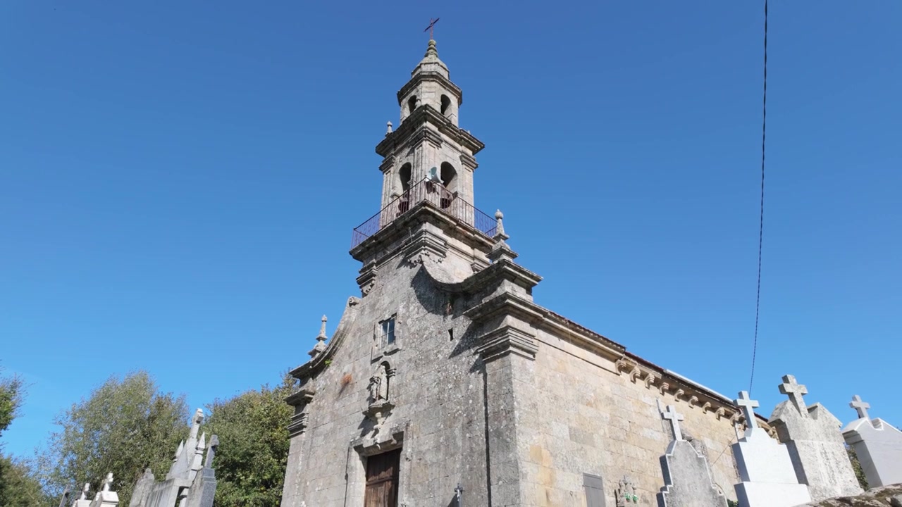 Vista em ângulo baixo da igreja de San Martiño de Porqueira em Ourense, Galiza, destacando sua fachada histórica de pedra, campanário e arquitetura católica tradicional sob um céu azul claro.