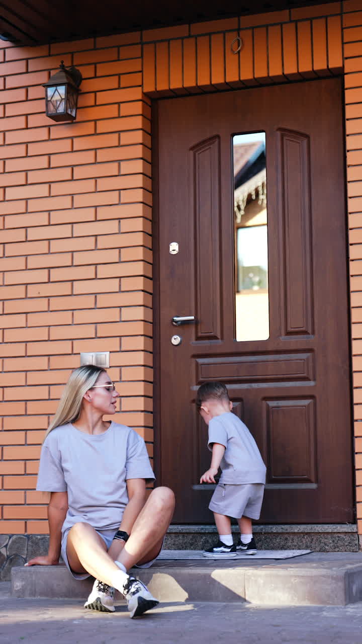 Mom and child outdoors. A mother relaxes on steps while her child plays nearby, both wearing matching gray outfits on a sunny day