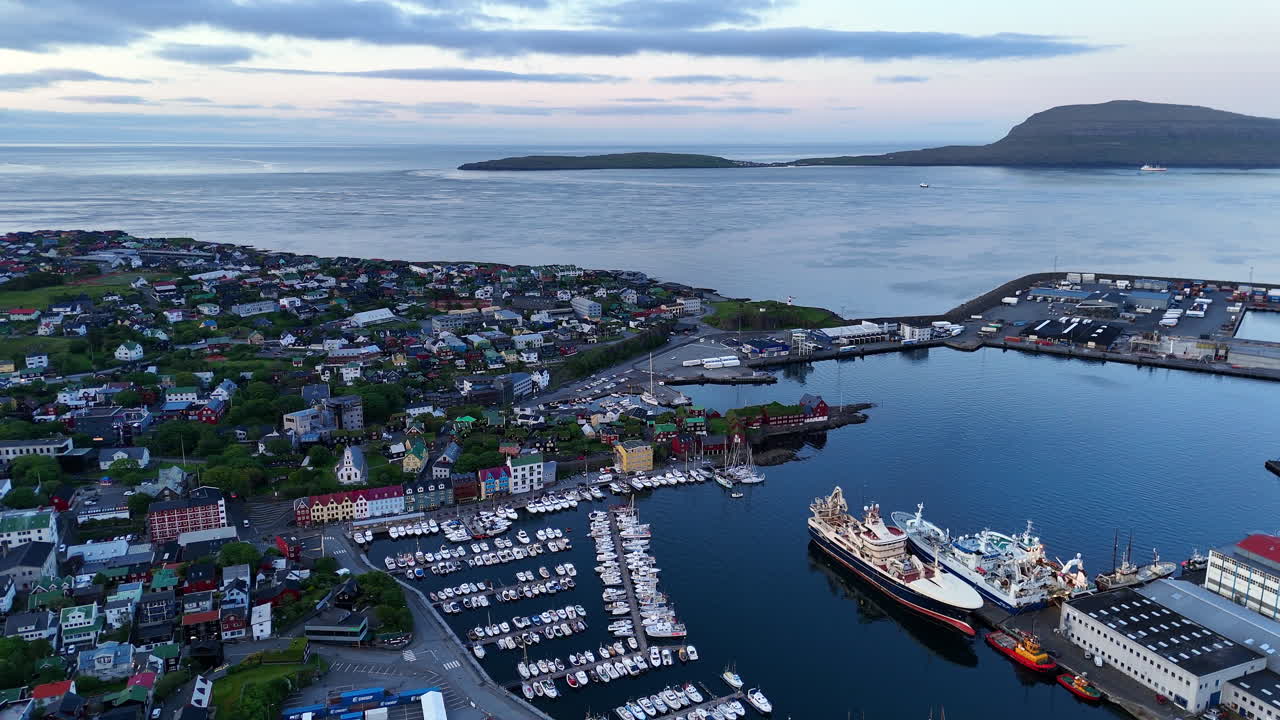 Cinematic aerial view of colorful village houses along a fjord in the Faroe Islands, surrounded by dramatic green mountains, black sand beach, and misty coastal scenery