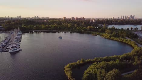 aerial sunset descending shot of sailboat and marina yacht club dock in lake bay surrounded by windy green trees with city buildings skyline in background in toronto ontario canada