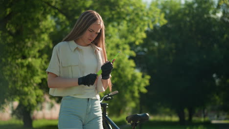 young blonde woman stands outdoors, concentrating as she struggles to adjust green air pump nozzle in her gloved hands, framed by lush trees and sunlight