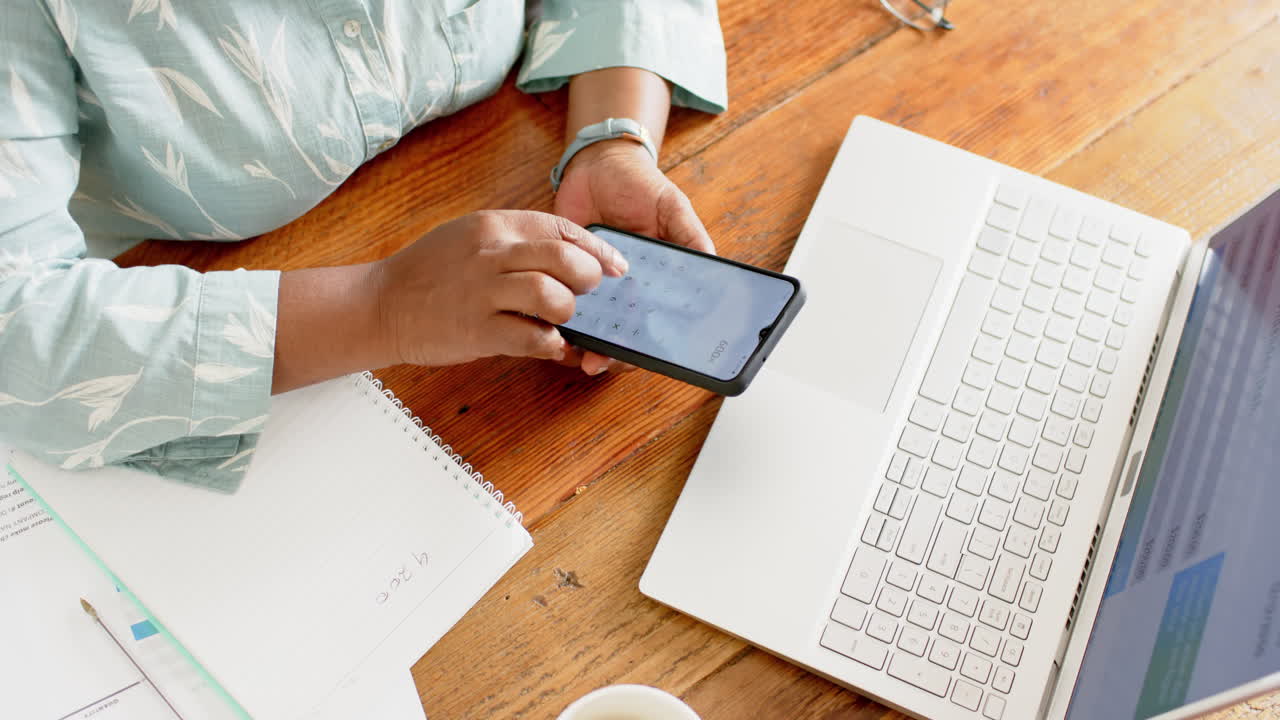 mujer afroamericana de edad avanzada haciendo papeleo, usando portátil, teléfono inteligente en una habitación soleada, cámara lenta