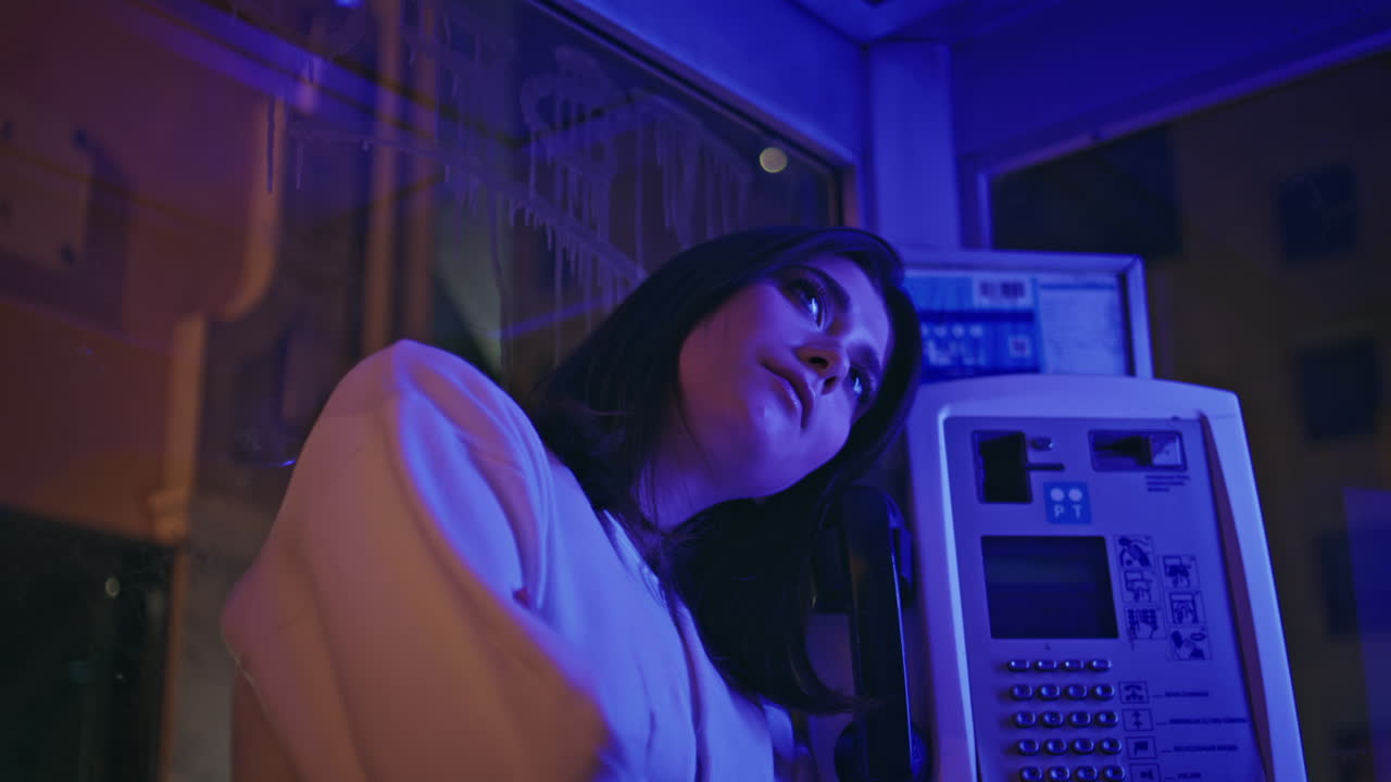 Primo piano: ragazza illuminata al neon che pensa in una cabina telefonica solitaria di notte. Donna appoggiata al telefono.