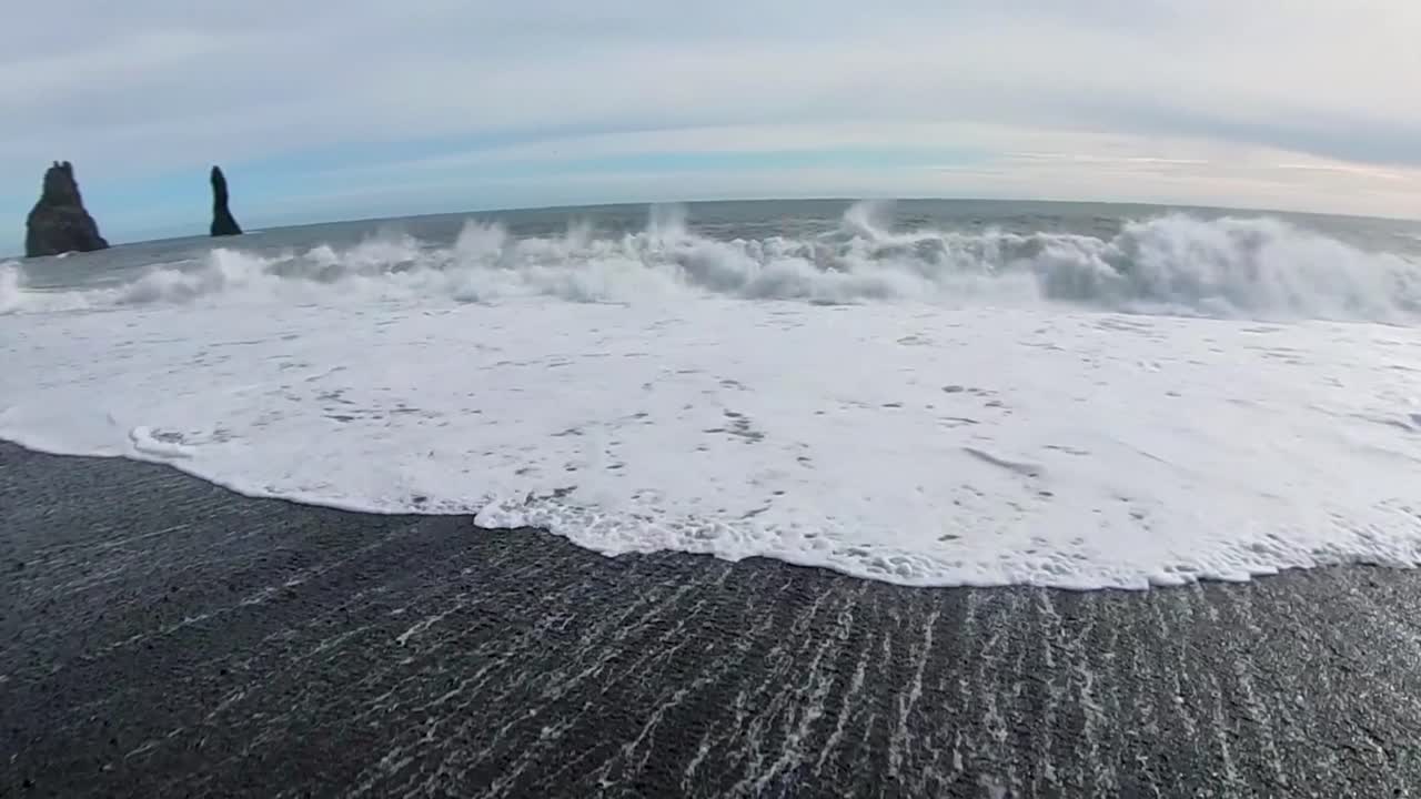 камера движется с жидкостью, фиксируя ритмический танец волн под облачным небом, черный песчаный пляж