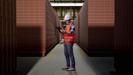 full body side view of asian male engineer with safety helmet working on a tablet while standing with red brick packed in stacks are stored