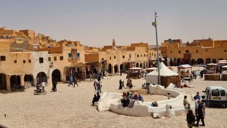 View on main square of Saharan oasis town with people passing and historical clay houses in the background on sunny and hot day in Ghardaia Algeria
