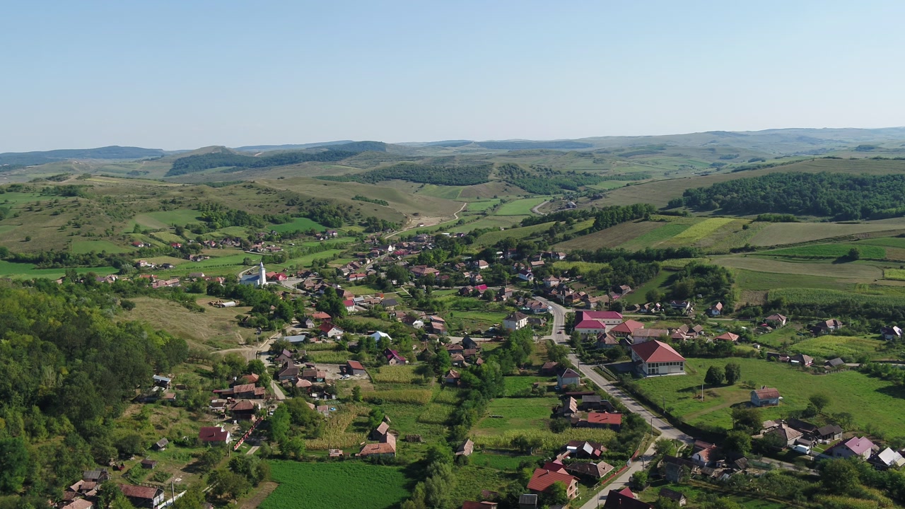 A static, high-angle aerial view of a rural Romanian village. A church stands at the center, surrounded by houses, green rolling hills, and pastures on a sunny summer day.