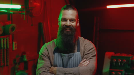 portrait shot of caucasian male welder with long beard and wearing apron, crossing hands and looking at camera in workshop