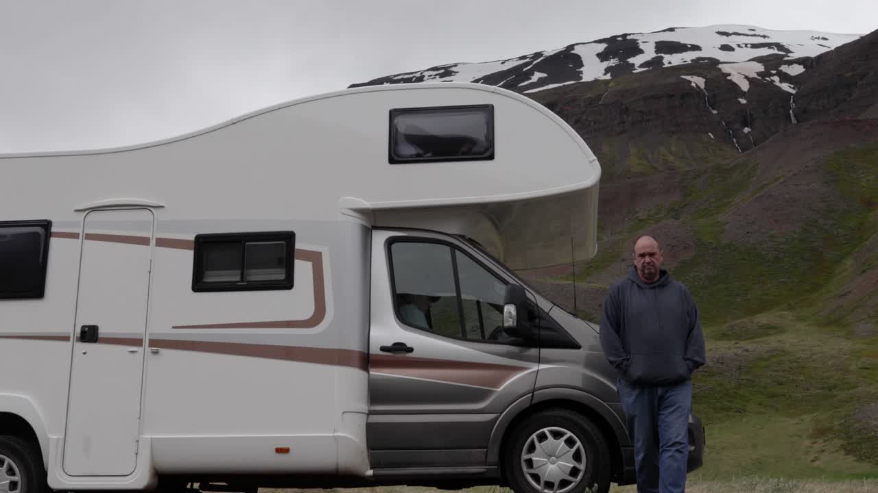 Uomo che cammina da un camper vicino alla telecamera con una montagna sullo sfondo