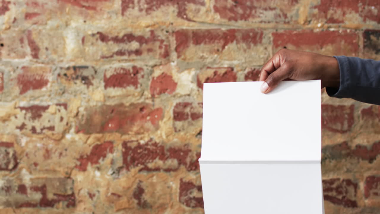 Video of hand of african american man holding book with blank pages, copy space over brick wall