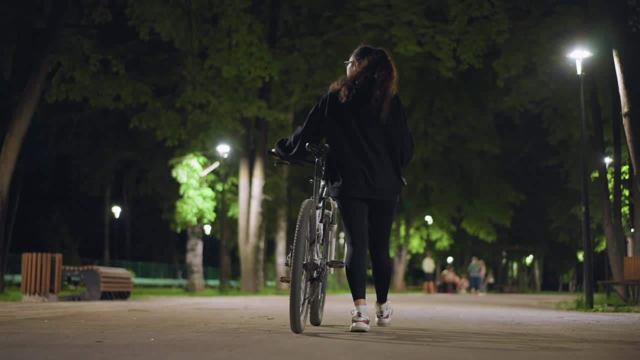 Quiet Night Scene With Cyclist And Bench, Calm Urban Setting Featuring Woman With Bicycle Under Light, Serene Nighttime Environment Depicting Woman Pausing Beside Her Bicycle Near Bench