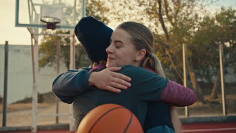 Two-happy-blonde-girls-in-sportswear-after-playing-basketball-give-each-other-high-fives-on-the-red-court-on-the-street-in-the-morning.-Happy-end-of-training-and-victory-after-playing-basketball-in-summer