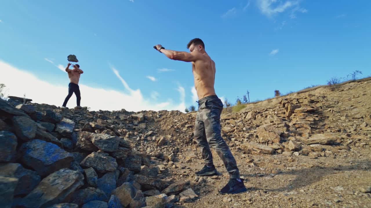 Strength sportsmen training on rocky background. One athlete breaking rocks with a heavy hammer while another man throwing stones. Slow motion.