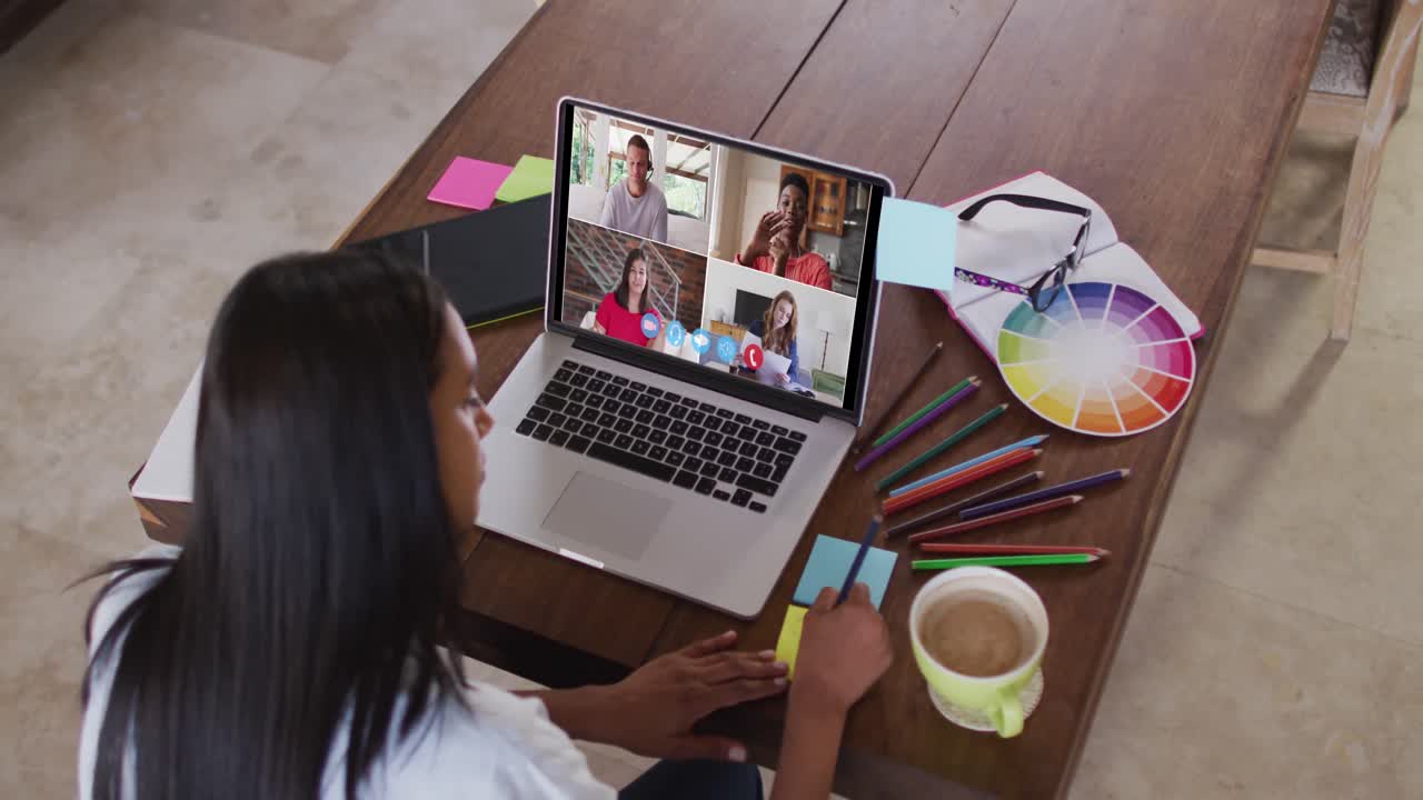 Mujer caucásica utilizando un portátil en una videollamada con colegas y tomando notas, trabajando desde casa.