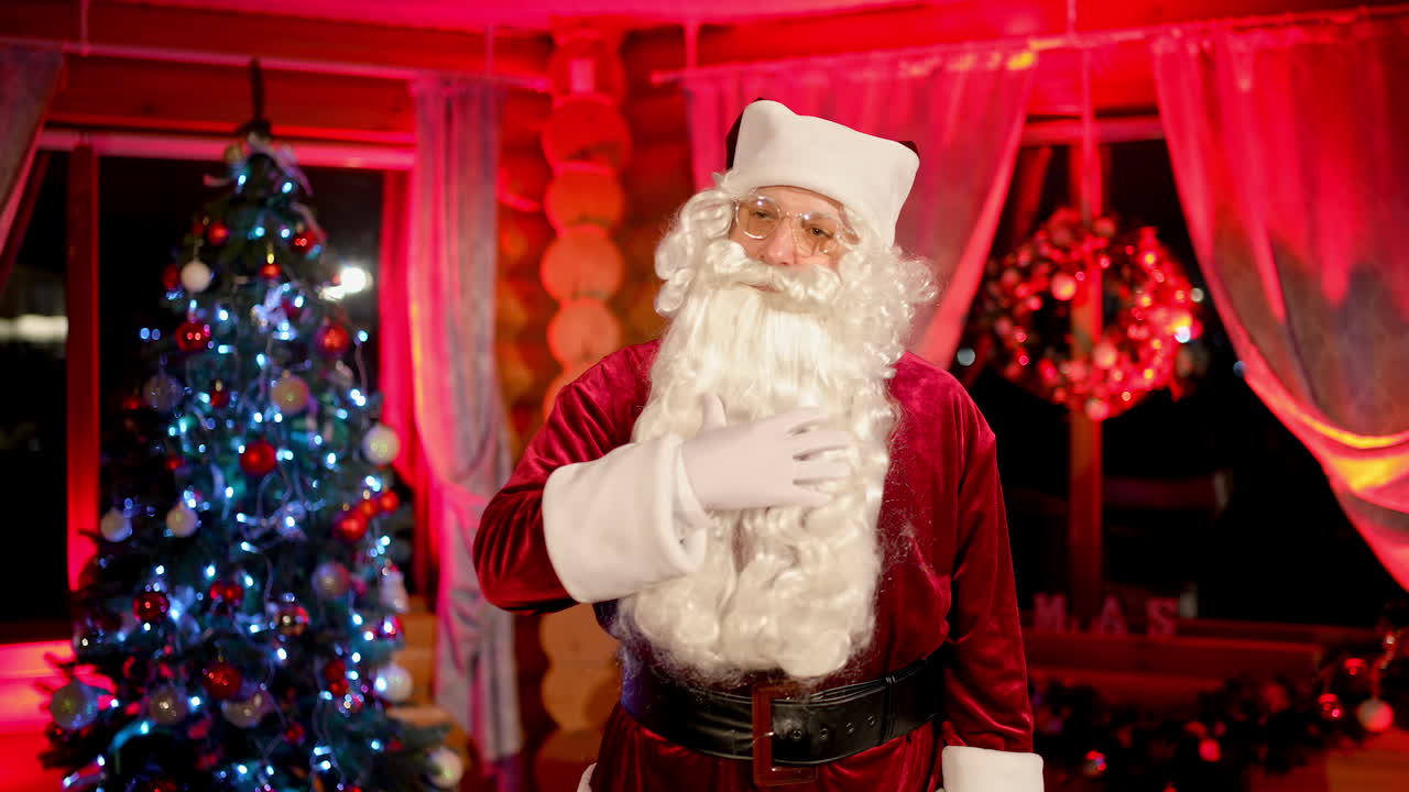Portrait of a senior man in santa claus costume indoors. Serious Santa with white beard standing in apartment on christmas tree background. Christmas spirit.