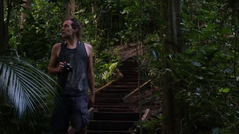 static shot of a tourist with camera in hand in the middle of a densely overgrown forest while walking down a wooden staircase and observing the beauty of nature on his journey through the jungle