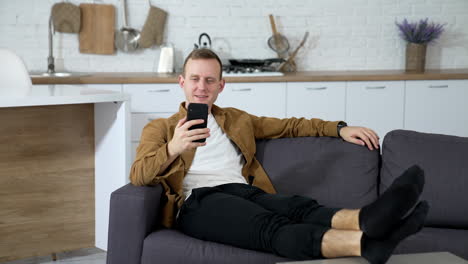 portrait of young man resting on sofa with a phone. guy is laying on a couch in the kitchen and uses mobile phone. student studying online through phone at home.