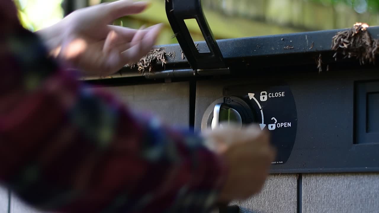 Beautifully shot alternate angle close up of a padlock and latch on a shed with hands reaching in to use a key to unlock and remove the lock and raise the lid, with stylish bokeh