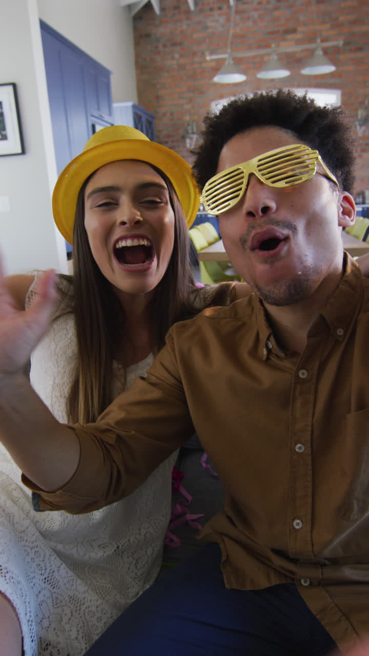video vertical de una pareja diversa celebrando con sombrero de fiesta y gafas haciendo una llamada de video en casa