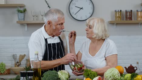 senior couple in kitchen. grandmother and grandfather feeding each other with raw vegetable salad