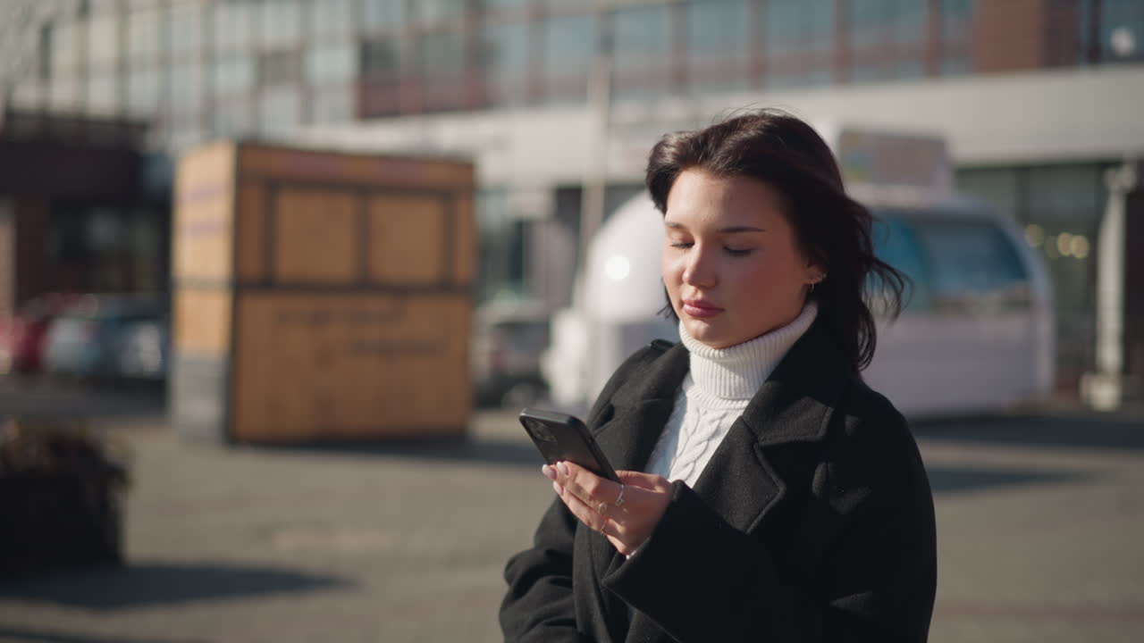 Elegant woman walking outdoors in black coat and white turtleneck, deeply focused on her smartphone while modern urban architecture reflects sunlight in the background