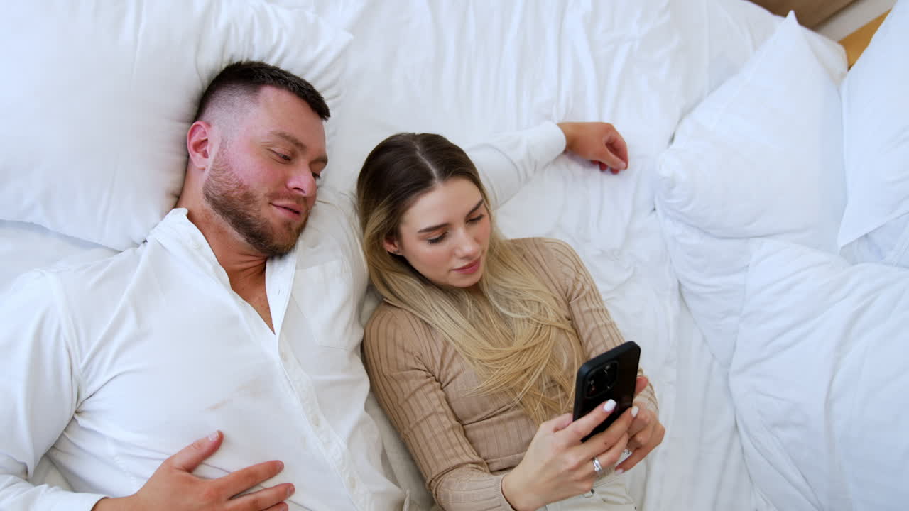 Caucasian couple lies on the bed together. Attractive woman is taking selfie of herself and her man. Top view.