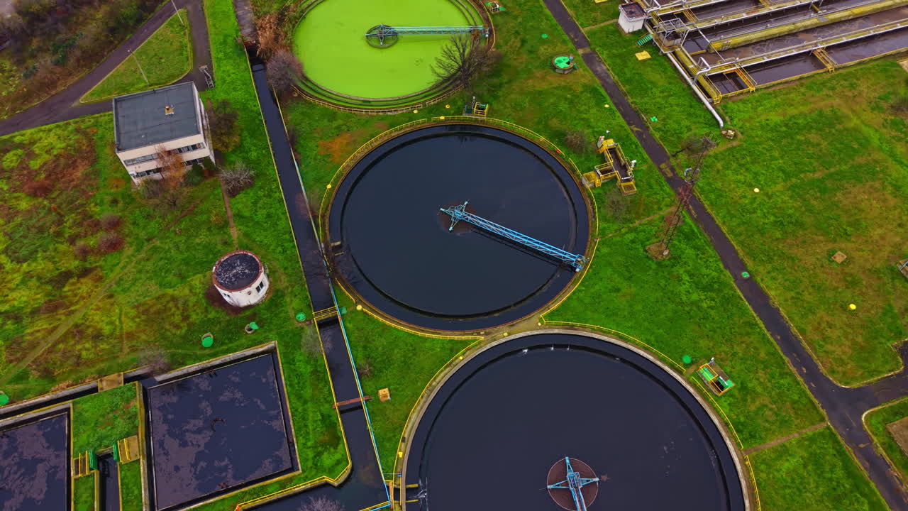 Wastewater treatment facility shows large circular tanks, green grass, and surrounding structures during daytime