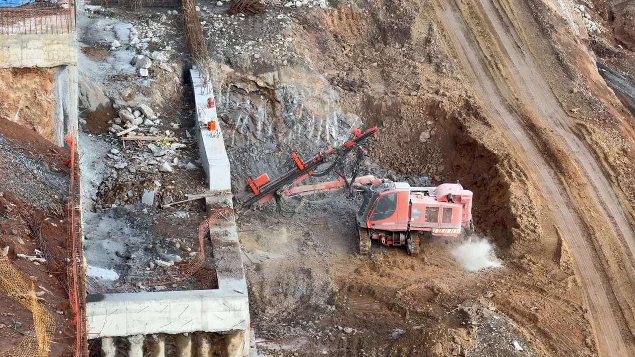 A large, tracked orange drilling rig is boring into a rock surface on a hillside or roadside for slope stabilization purposes. Construction, geology, rock ground, and road construction infrastructure.