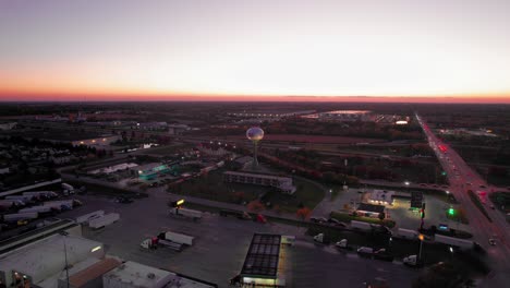 Aerial-dusk-view-of-Monee,-IL:-glowing-sunset-over-urban-sprawl,-roads,-and-water-tower