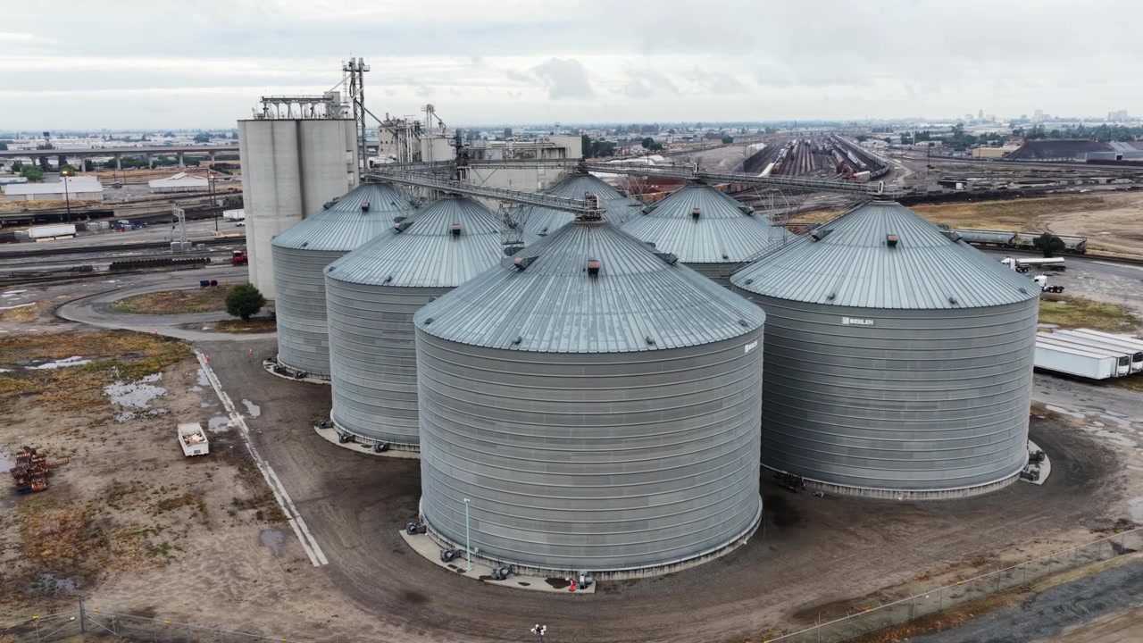 Eine Gruppe von Stahlgetreidesilos erhebt sich auf einem industriellen Lagergelände nahe Fresno, Kalifornien, USA, umgeben von Fördertürmen und einem Kiesplatz unter einem dichten, bedeckten Himmel. Luftaufnahme mit Drohne. Weitwinkelaufnahme