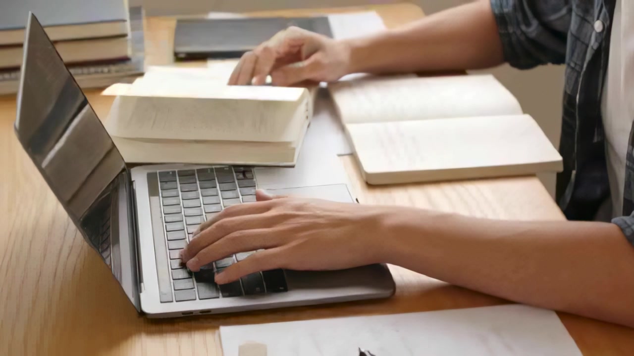 Mann i rutete skjorte taster med venstre hånd på tastene mens høyre hånd svever, blar gjennom laptop eller bøker under research ved pulten.