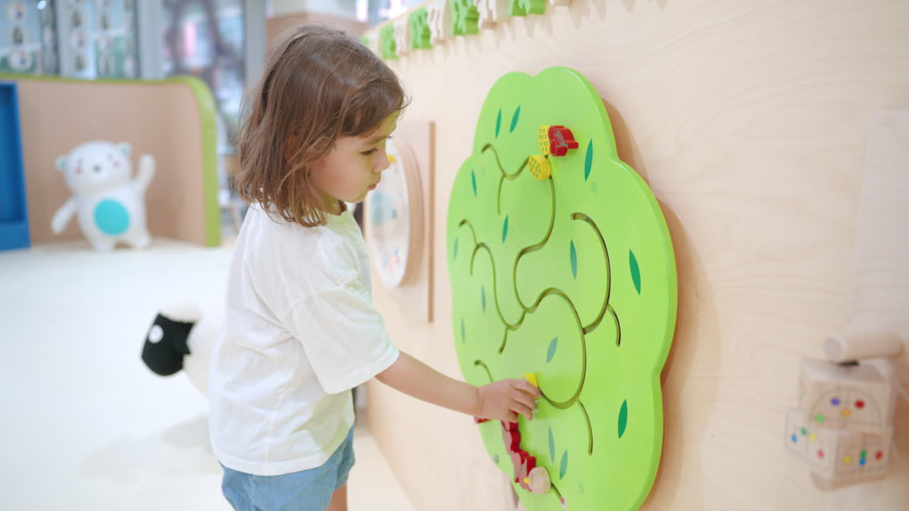 유아 소녀가 놀이실 내부의 나무 놀이 벽을 따라 슬라이딩 과일을 움직여서 놀고 있습니다.