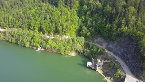 Toma-Aérea-Hacia-Abajo-De-La-Carretera-Junto-Al-Embalse-Del-Lago-Con-árboles-Junto-Al-Frente-De-Agua