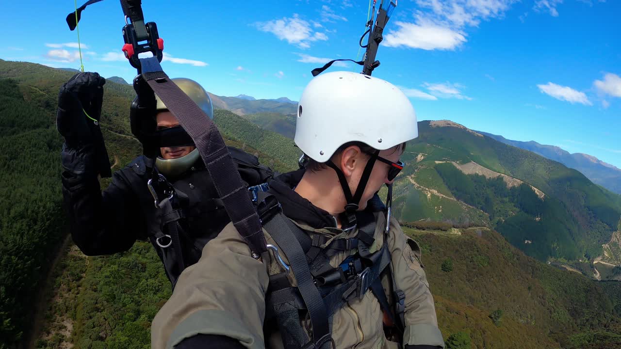 Father and son in tandem paragliding over forested valleys in Tasman, New Zealand on clear day, selfie pov