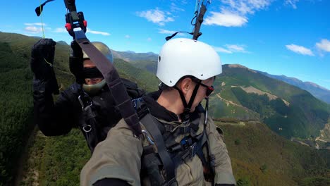 father and son in tandem paragliding over forested valleys in tasman, new zealand on clear day, selfie pov