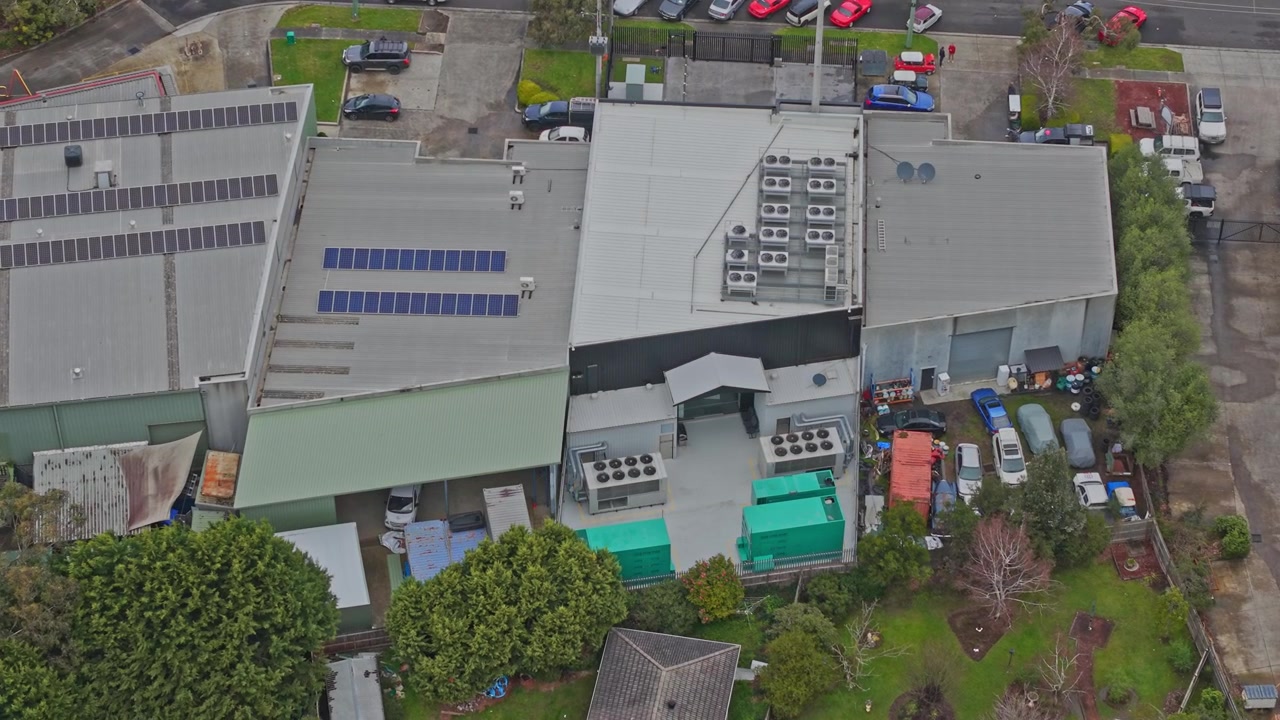 Dynamiske luftoptagelser af et moderne datacenterkompleks med HVAC-systemer på taget, solpaneler og sikker infrastruktur. Overskyet belysning, med omgivende grønne områder og synlige parkeringsområder