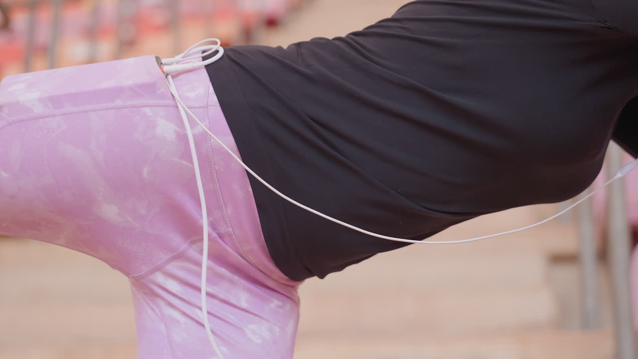 Jeans rosa e catena sui fianchi, appoggiata sulle gradinate, primo piano di top nero che mostra busto e curve, atmosfera di moda casual da strada, sedili dello stadio all'aperto illuminati dal sole, catena metallica appesa al passante della cintura, atteggiamento moderno e sicuro.
