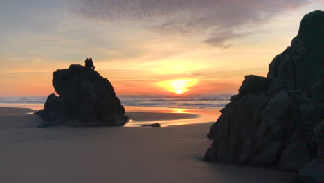 two people sitting on a tall rock at devil's kitchen, which is a part of bandon beach state park at the oregon coast, enjoying a peaceful and beautiful sunset