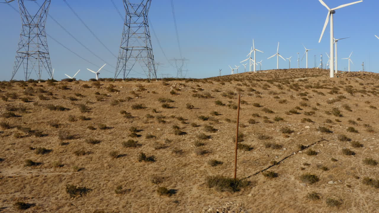drone volando hacia atrás cuesta abajo mientras las turbinas eólicas y las líneas de transmisión de alto voltaje desaparecen detrás de la colina cerca de palm springs en el desierto de mojave, california, ee.uu.