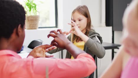 Diverse-female-teacher-and-happy-schoolchildren-at-desks-learning-sign-language-in-school-classroom