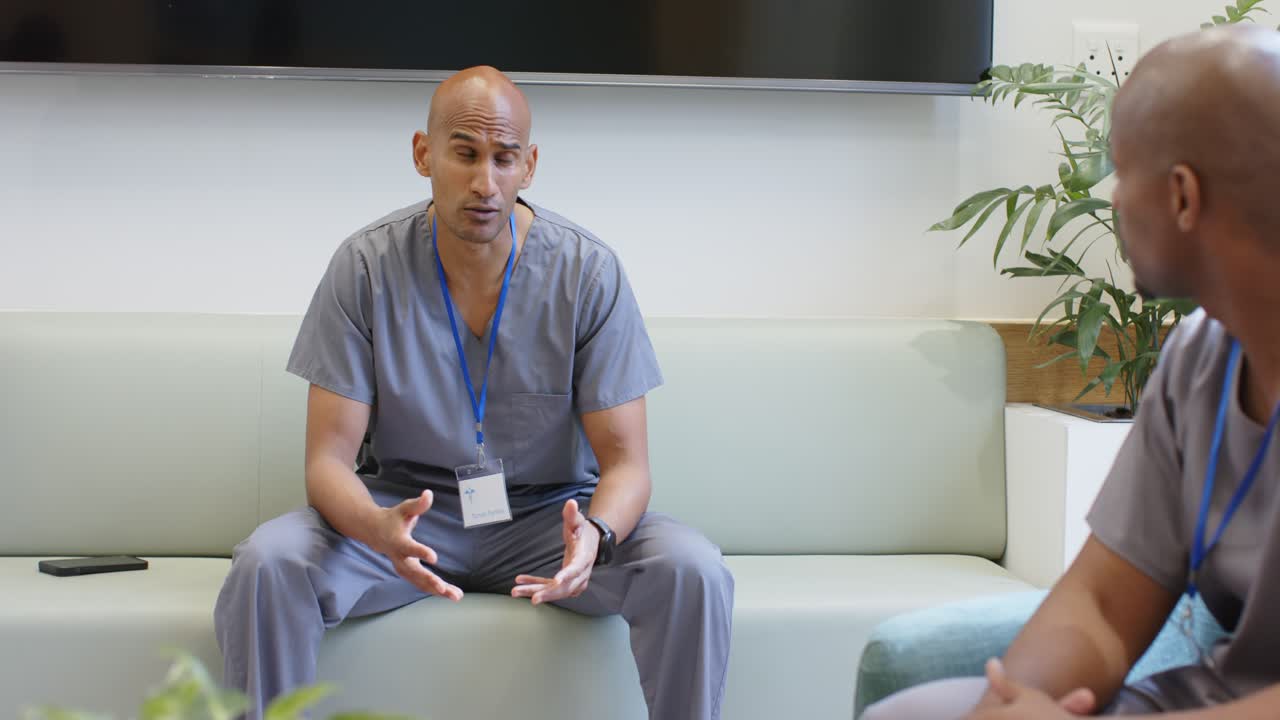 Un membro del personale medico maschile in scrub si appoggia e parla, con una tazza, nella sala relax, dopo un cambio di inquadratura.