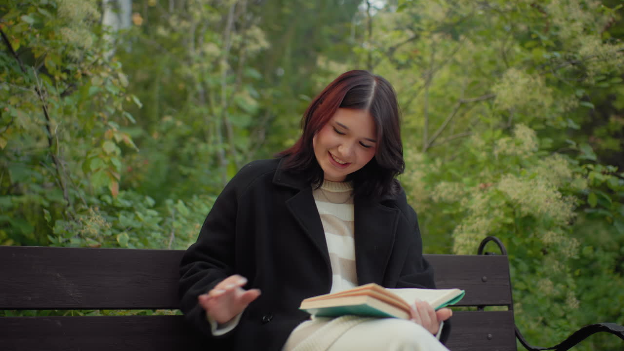 Serene woman seated on bench, reading novel, flipping pages with gentle smile, lifts cup of tea to sip then sets it beside her, calm autumn arboretum background, soft greenery and quiet mood