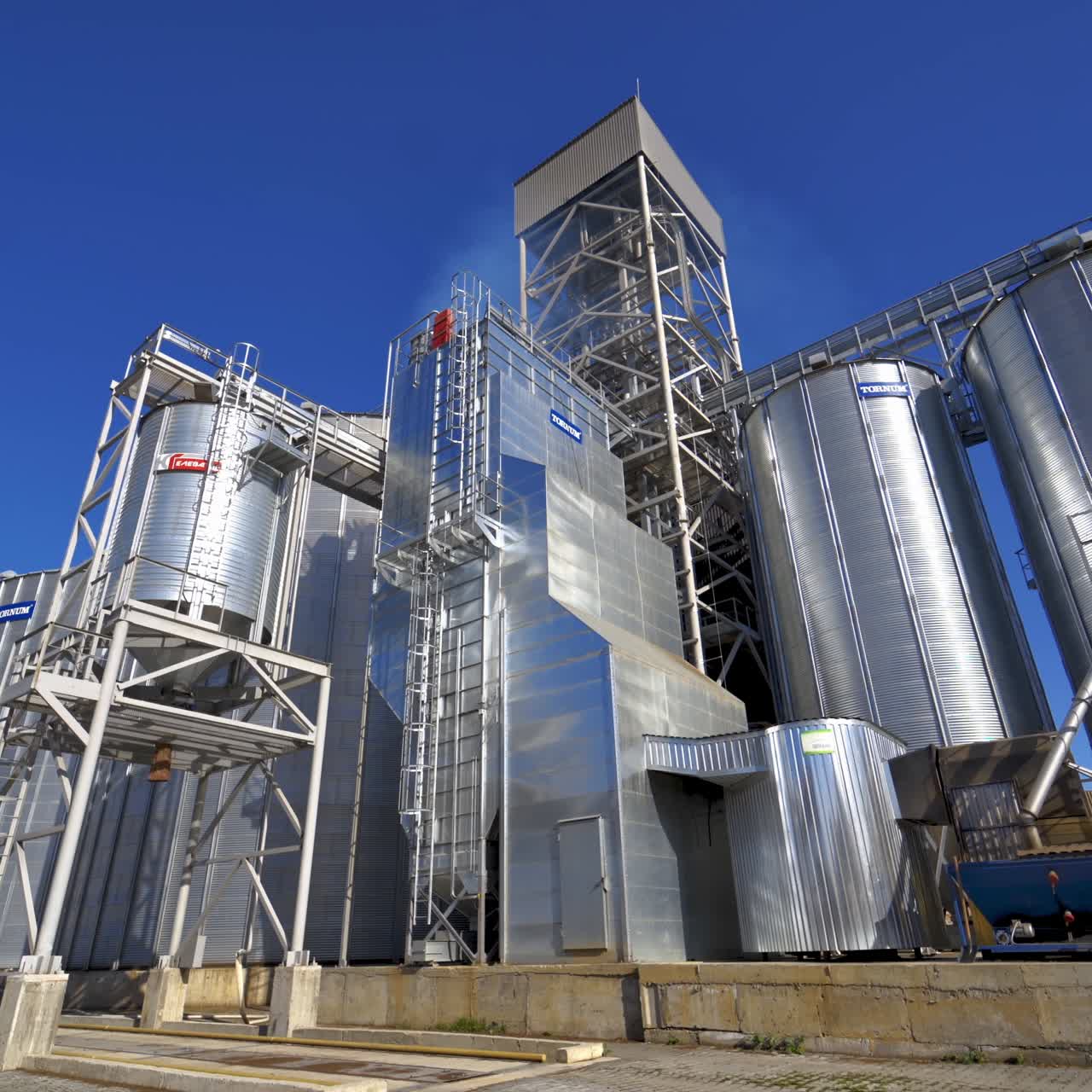 Modern plant for agribusiness. Silver grain elevators on blue sky background. Granary on field. Industrial factory with large storage bins at sunlight.