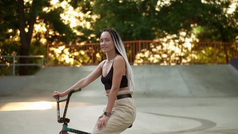 Portrait-of-a-beautiful-girl-with-black-and-white-dreadlocks-sitting-on-the-bike-in-skatepark-and-smiling-to-the-camera