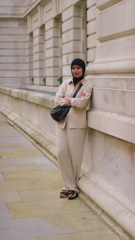 vertical video portrait of smiling muslim businesswoman wearing hijab and modern business suit standing and folding arms outside city office buildings 2