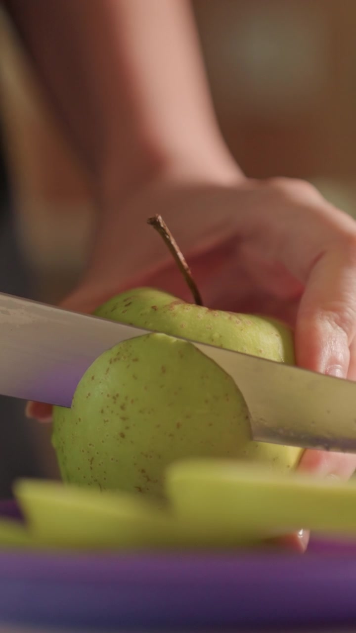 Primo piano di mani che sbucciano una mela verde. La buccia della mela cade mentre le fette si accumulano sotto, rivelando la polpa interna. L'illuminazione calda degli interni enfatizza il dettagliato processo di preparazione del cibo