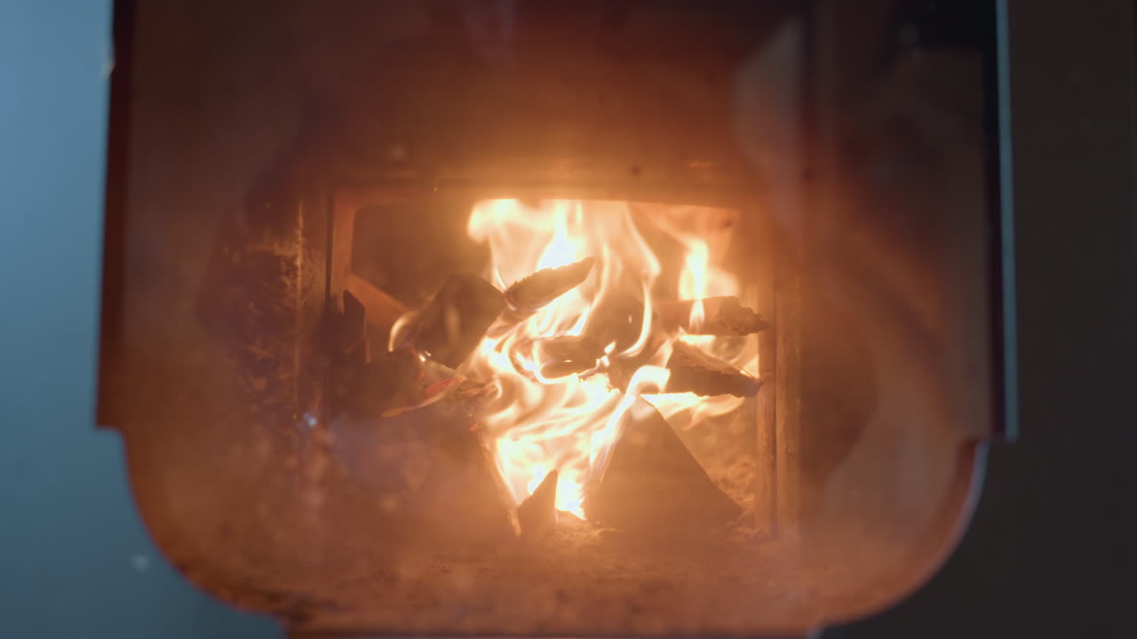 Close up of firewood burning inside enclosed fireplace, bright flames flicker and dance around stacked wooden logs, casting warm glow through slightly smudged glass door in indoor rustic setting