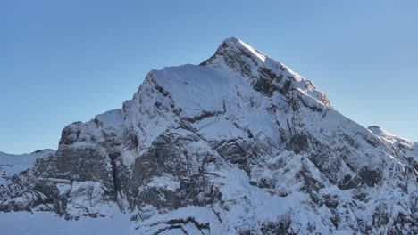 aerial view of fronalpstock in kanton glarus, switzerland. snow blankets the steep rocky slopes under a clear blue sky, showcasing the rugged beauty of the alpine peak in winter.