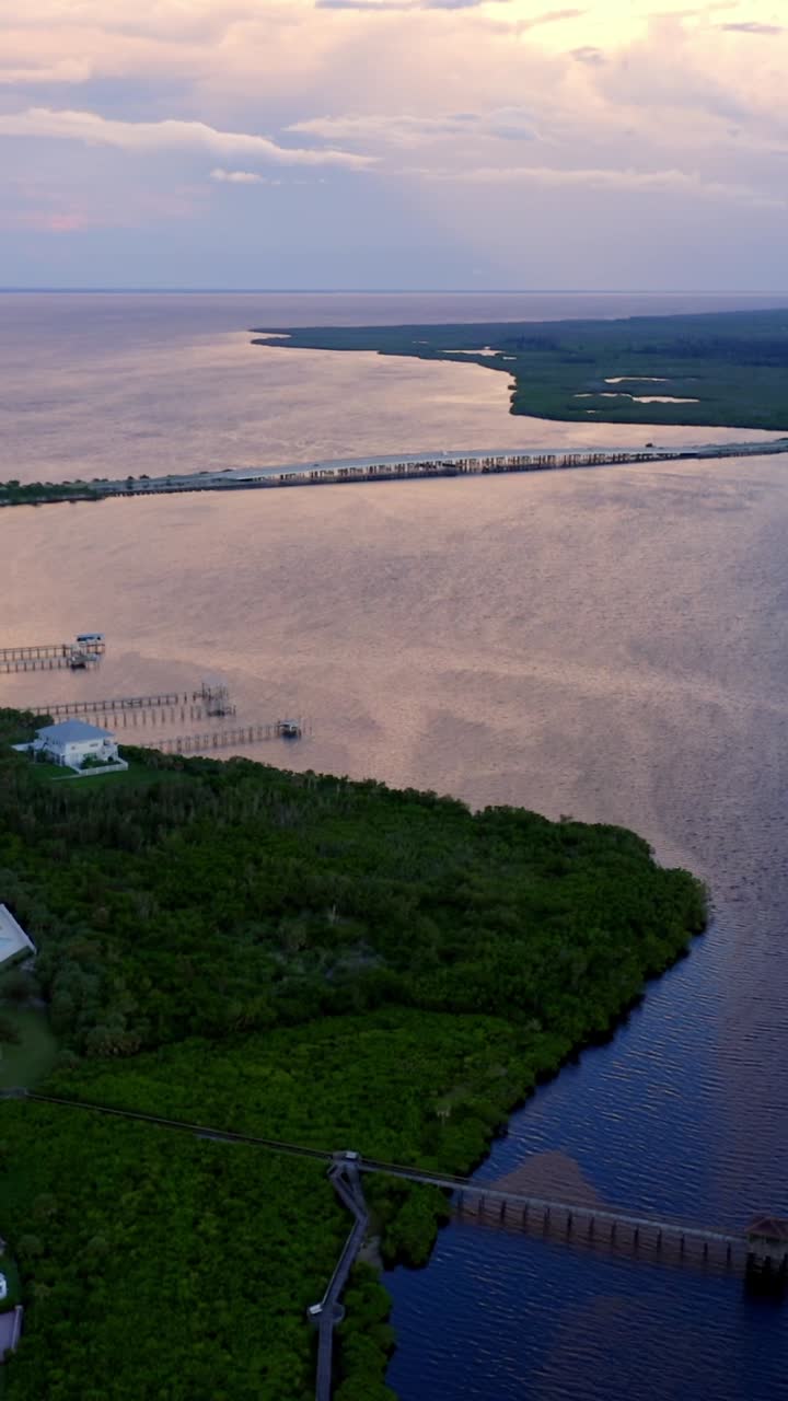 Warm light from the setting sun reflects across calm bay waters beside a coastal neighborhood of red-roofed homes, green mangroves, and a distant bridge fading into the horizon